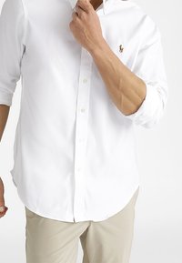 Bílá košile s dlouhými rukávy, slim fit, s malým vyšitým logem na levé hrudi, spárovaná se světle béžovými kalhotami.
