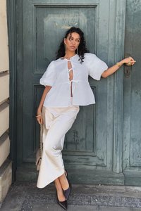Witte blouse met korte mouwen, knopen en uitsnijdontwerp, gecombineerd met een crèmekleurige satijnen rok en zwarte puntige hakken, voor een groene deur.