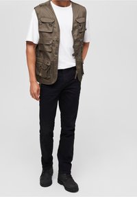 Khaki Weste mit mehreren Taschen, Reißverschlussverschluss, getragen über einem weißen T-Shirt. Kombiniert mit schwarzen Hosen und schwarzen, robusten Schuhen.