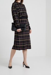 Suéter de punto con estampado de cuadros negro con líneas rosas y beige, combinado con una falda a juego. Sosteniendo un bolso de mano negro, lleva zapatos de tacón negros.