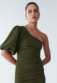Oliivinvihreä, yhdellä olkapäällä oleva mekko, jossa on teksturoitu yläosa. Sisältää puffihihan ja laskostettuja muotoiluosia, luoden istuvan siluetin.