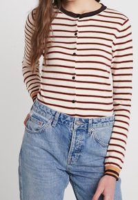 Gestreifter Cardigan in Creme und Braun mit Rundhalsausschnitt, Knopfleiste und kontrastierenden gerippten Bündchen. Kombiniert mit hoch taillierten blauen Jeans.