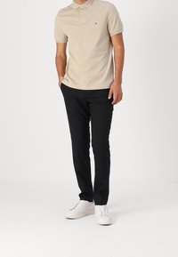 Beige polo tričko s krátkým rukávem a límečkem, spárované s černými kalhotami a bílými teniskami. Obsahuje vyšívané logo na hrudi.