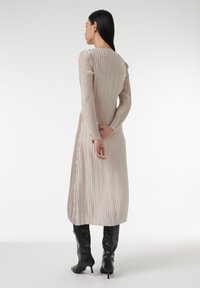 Robe longue plissée de couleur beige avec des manches longues, présentant une texture lisse et un design épuré. Associée à des bottes noires montantes.