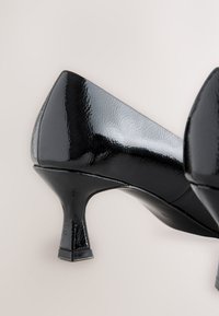 Crne salonke od lakirane kože s izduženim vrhom i niskom četvrtastom potpeticom. Površina je sjajna s teksturiranim finišem.