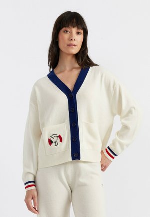 Cremfarbener Cardigan mit marineblauem Saum, Knopfverschluss und zwei Taschen mit einem gestickten Snoopy-Motiv. Gerippte Bündchen mit roten und marineblauen Streifen.