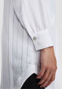 Vit skjorta med vertikala ränder, långa ballongsärmar, knappade manschetter med en klar sten detalj, och en asymmetrisk nederkant.