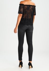 Schwarzes Spitzen-Oberteil mit schulterfreier Gestaltung, floralem Muster, halbtransparenter Stoff und gewelltem Saum, kombiniert mit dunklen Jeans und schwarzen Absätzen.