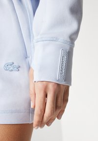 Hanorac albastru deschis, realizat dintr-un material neted, cu manșete cu textură ribbed și un patch cu logo texturat pe mânecă. Prim-plan cu o mână sprijinită de articol.
