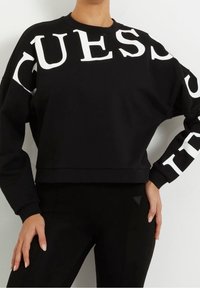 Schwarzes, kurzes Sweatshirt mit großen weißen "GUESS"-Buchstaben auf der Vorderseite und den Ärmeln. Der Stoff ist glatt und hat gerippte Bündchen.