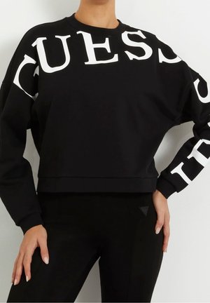 Zwarte cropped sweatshirt met grote witte "GUESS" letters over de voorkant en mouwen. De stof is glad met geribbelde manchetten.