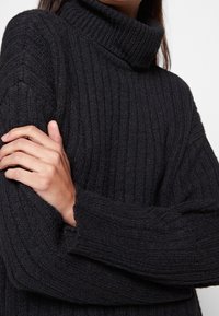 Czarny sweter z golfem, prążkowany, z długimi rękawami i luźnym krojem. Tekstura wydaje się pleciona i miękka, podkreślając ciepło i komfort.