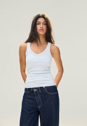 Jeune femme aux longs cheveux bruns portant un débardeur blanc et un jean bleu foncé, debout avec les mains derrière le dos.