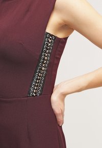 Robe bordeaux sans manches avec un détail en lacets noirs sur le côté, portée par une personne avec la main posée sur la hanche.