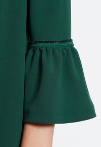 Grön klänning med utsvängd ärm, med volang nederkant och ett svart accentband vid cuffen. Slät tygtextur.