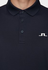 Mörkblå polo skjorta i ett mjukt tyg, med klassisk krage, tre knappar och en liten vit logotyp på vänster sida.