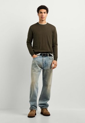 Junger Mann steht, trägt dunkelgrünen Pullover, hell ausgewaschene Jeans mit schwarzem Gürtel, braune Schuhe und eine Hand in der Tasche vor einfarbigem Hintergrund.