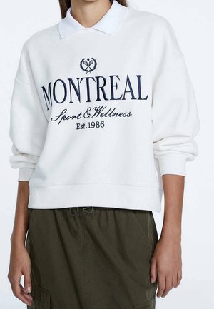 Biała bluza z długim rękawem z granatowym napisem „MONTREAL Sport & Wellness Est. 1986”, noszona z oliwkowozielonymi spodniami z wysokim stanem.