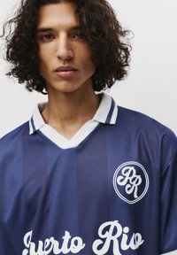 Maillot de sport bleu marine avec des rayures verticales, col blanc, et lettres "Puerto Rio". Présente un logo circulaire avec les initiales "PR".