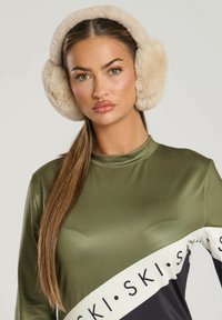 Faux pels ørevarmere i beige, blød tekstur, rund form. Bæres med en grøn langærmet top med et kontrasterende sort-hvidt grafik.