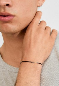 Helmestruktuuriga käevõru, millel on mustad, hallid ja kuldsed helmed õhukesel elastsel nööril, kantud kergelt tumeda naha tooniga randmel.