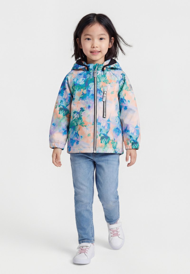 Νέα κοπέλα που περπατά, φορώντας πολύχρωμο φλοράλ μπουφάν με κουκούλα, ανοιχτό μπλε τζιν και λευκά αθλητικά παπούτσια με ροζ κορδόνια.