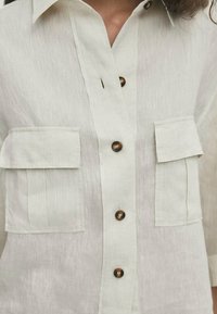 Linen button-up shirt in light beige with two front pockets, dark brown buttons, and a classic collar design.