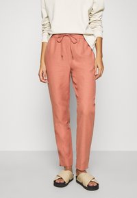 Volné hnědé kalhoty v terakotové barvě s elastickým pasem a šňůrkou. Hladká tkanina; zúžený střih nohavic. Nosené s krémovým svetrem a sandály.