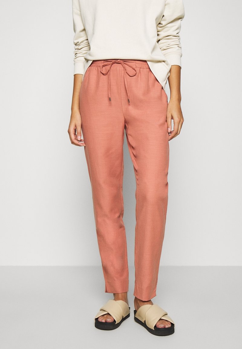 Volné hnědé kalhoty v terakotové barvě s elastickým pasem a šňůrkou. Hladká tkanina; zúžený střih nohavic. Nosené s krémovým svetrem a sandály.