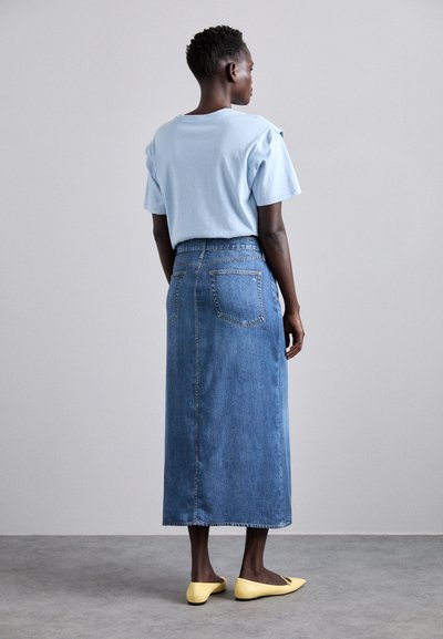 T-shirt bleu clair à manches courtes associé à une longue jupe en denim. La jupe possède deux poches arrière et une coupe droite, portée avec des ballerines jaunes.
