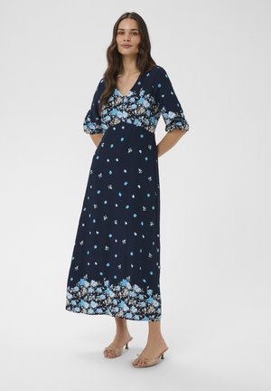Femme portant une robe midi bleu marine avec des motifs floraux bleus et blancs, des manches ballon longueur coude et des escarpins ouverts beige.