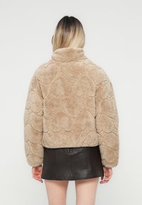 Kvinde med ombre hår iført en beige struktureret falsk pelsjakke og en sort læder mini nederdel, stående med ryggen til kameraet.