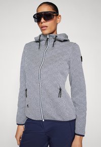 Giacca zip-up a motivi in bianco e nero con cappuccio, dotata di due tasche laterali e accenti neri. Tessuto testurizzato con un design geometrico.
