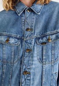 Jeansjacka i ljusblått med två bröstfickor, bruna knappar med Lee-logotypen och kontrasterande gula sömmar.