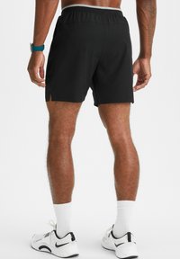 Schwarze Sportshorts mit einem grauen elastischen Bund, gespaltenem Saumdesign und glatter Textur. Getragen mit weißen Sportschuhen und Socken.
