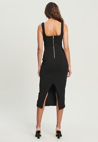 Femme se tenant de dos, portant une robe midi sans manches noire avec une fermeture éclair au dos et une fente centrale, assortie de talons noirs.