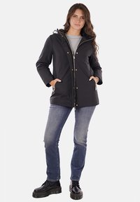 Giacca nera con cappuccio, chiusura con zip e bottoni a pressione, tasche laterali e accessori in tonalità dorata, indossata con jeans blu e stivali neri.