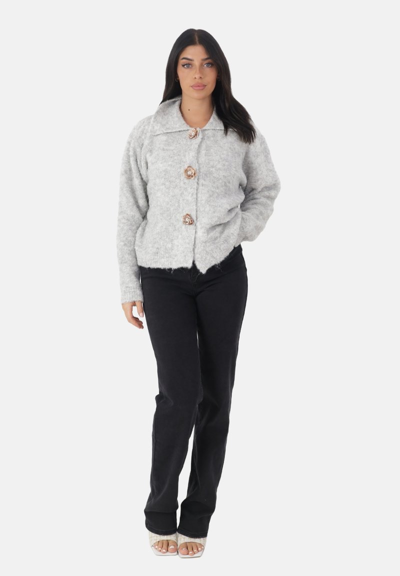 Szary, puszysty kardigan z kołnierzem i różowo-złotymi guzikami, w połączeniu z czarnymi spodniami. Modelka stoi z rękami w kieszeniach, nosząc sandały na obcasie z odkrytymi palcami.