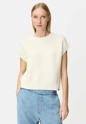 Blonde Frau mit weißem, kurzärmeligem Stricktop und hellblauen, hoch taillierten Jeans, die die Hände in den Taschen hat, neutraler Hintergrund.