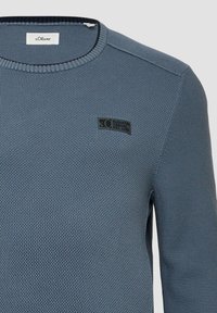 Hellblauer, strukturierten Pullover mit Rundhalsausschnitt, der ein kleines schwarzes Logopatch auf der linken Brust und Rippdetails am Kragen aufweist.