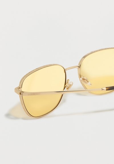 Γυαλιά ηλίου με χρυστό μεταλλικό σκελετό, φακούς με κίτρινη απόχρωση και ρυθμιζόμενα ρινοπέδια, που ακουμπούν σε λευκή επιφάνεια.