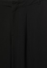 Primo piano di pantaloni in tessuto nero con cintura visibile, passanti per cintura e sottili pieghe frontali.