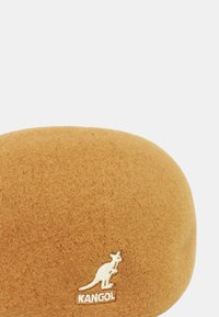 Beige vilten baret met een gladde textuur, voorzien van een witte geborduurde kangoeroe-logo en merknamen aan de onderkant. Ronde vorm.