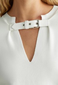 Gros plan sur un décolleté en V avec une bretelle en tissu blanc maintenue par une boucle en argent sur un haut blanc texturé.