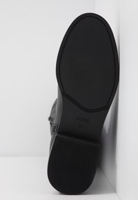 Semelle noire d'une botte de la marque Guess, taille 37, avec un talon texturé et une zone des orteils lisse, appuyée contre un fond d'angle blanc.