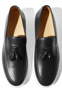 Scarpe slip-on in pelle nera con suole in gomma bianca, dotate di nappine decorative e una texture liscia. Dettagli del marchio visibili all'interno.