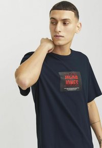 Námorně modré tričko s krátkým rukávem, které má obdélníkovou grafiku v červené a černé barvě s textem "JACK & JONES". Hladká textura látky, volný střih.