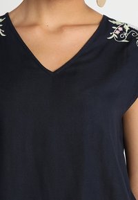 Mörkblå V-ringad topp med korta ärmar, prydd med lätt blomsterbroderi längs axlarna. Slät tyg med en mjuk textur.