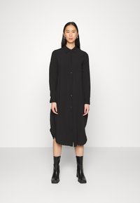 Vestido preto de botões, com mangas longas, colarinho e fendas laterais. Usado com botas pretas de salto até à metade da panturrilha. Textura suave.