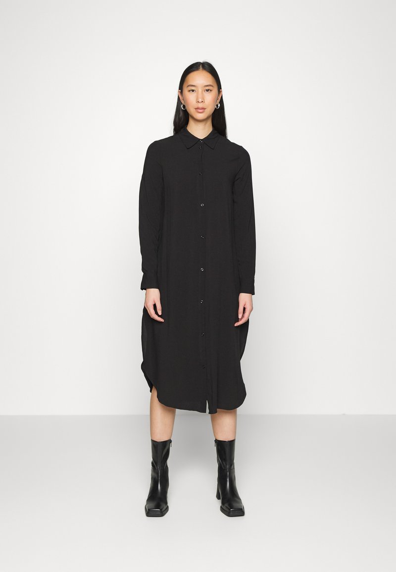Vestido preto de botões, com mangas longas, colarinho e fendas laterais. Usado com botas pretas de salto até à metade da panturrilha. Textura suave.